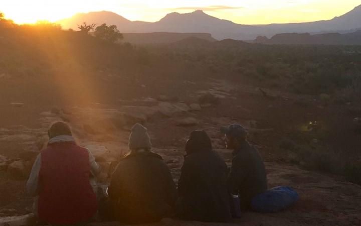 Three students sit facing away from the camera watching the sun rise or set in a desert landscape.
